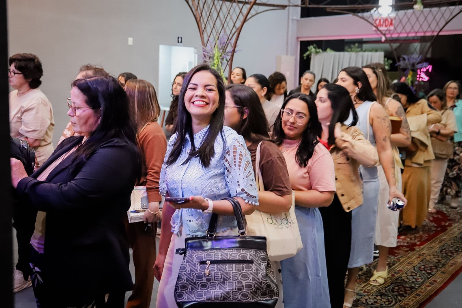 Mulheres chegando e se cumprimentando no espaço da conferência, com uma participante sorrindo para a câmera.