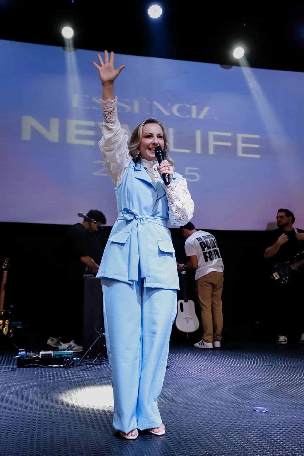 Preletora no palco com o microfone e uma das mãos levantada, diante do telão com o nome New Life.