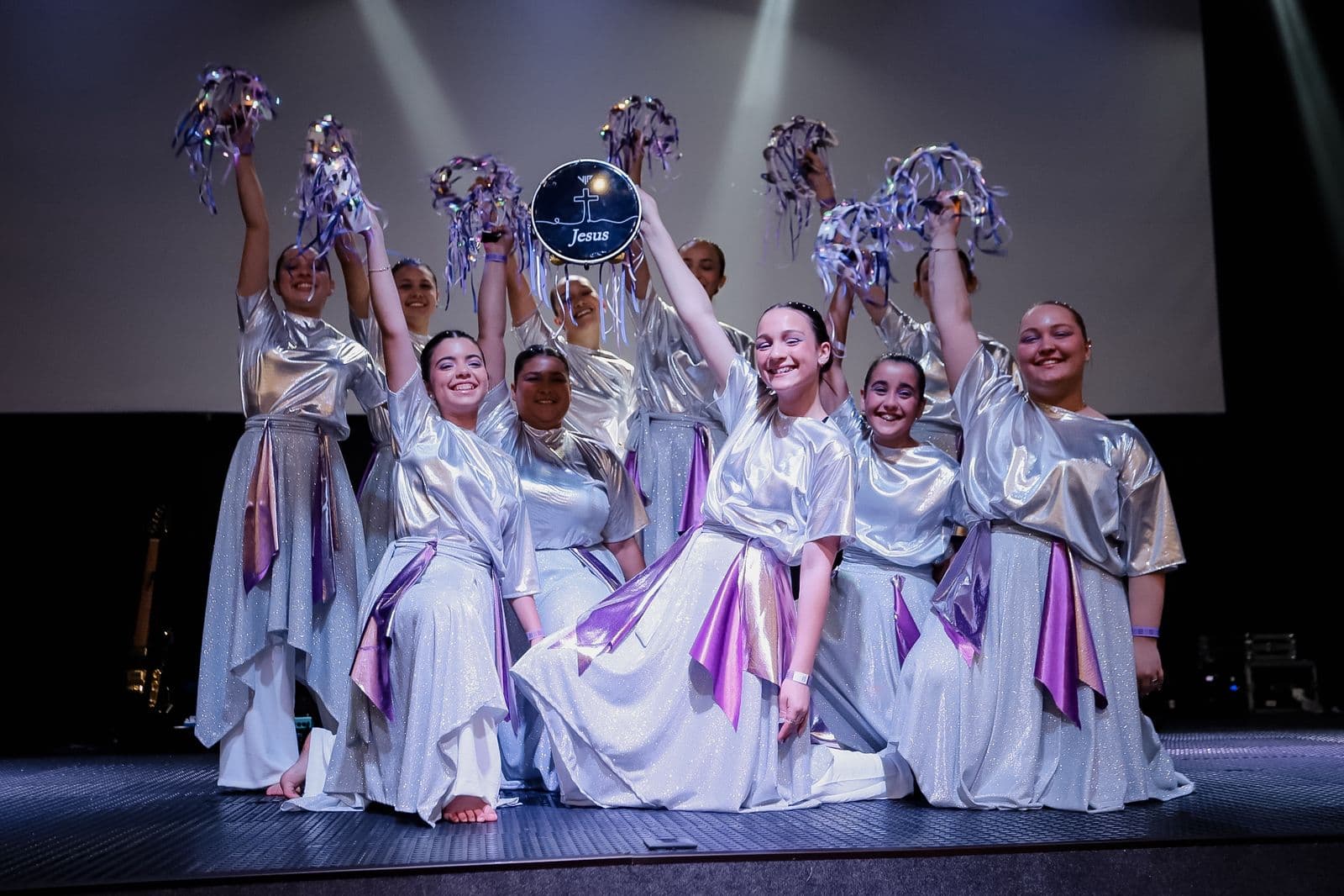 Grupo de mulheres em traje de dança prateado no palco, com os braços erguidos e um pandeiro escrito Jesus.