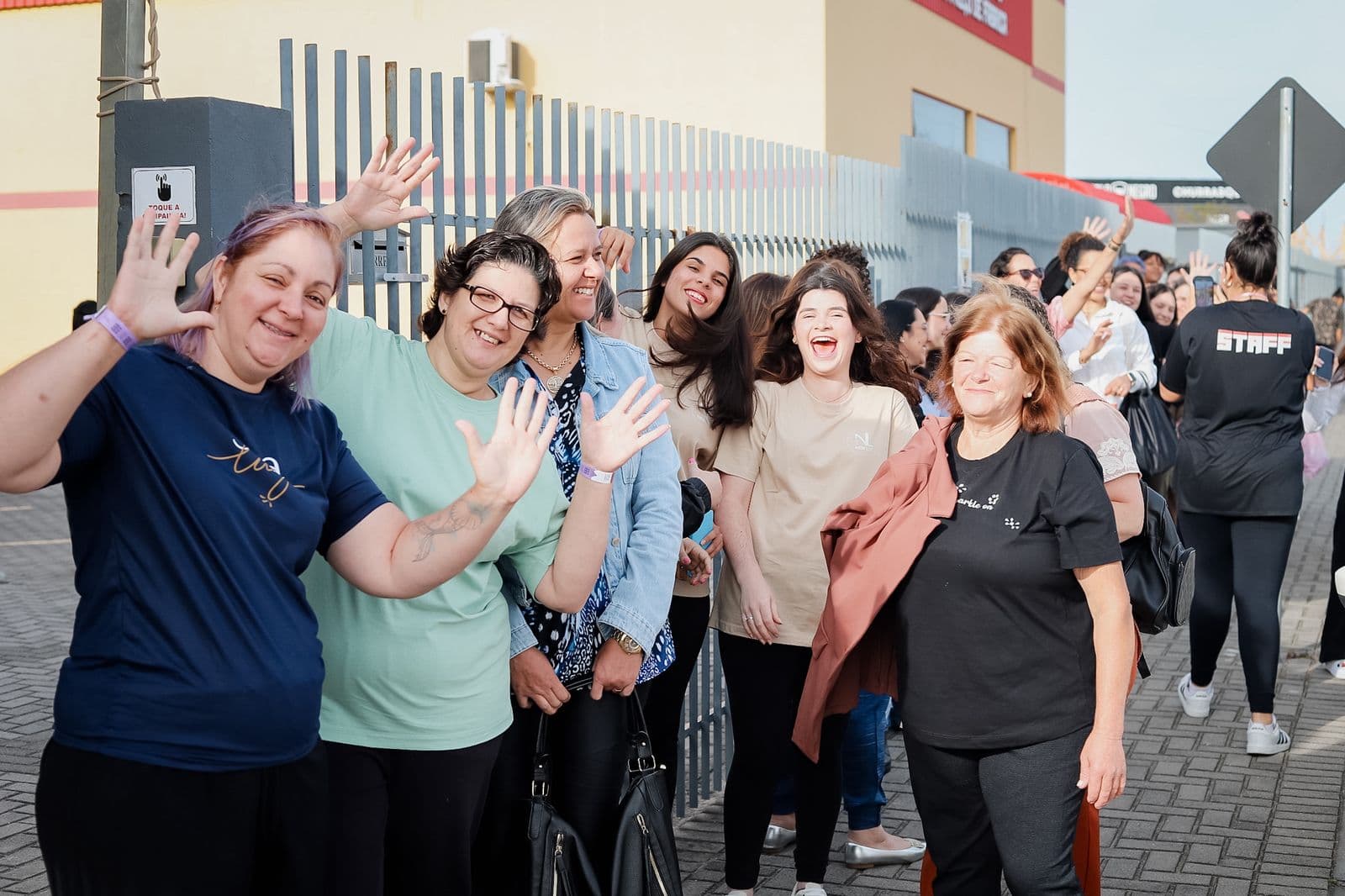Grupo de mulheres sorrindo e acenando na entrada do evento, em um dia de sol, na chegada da conferência.