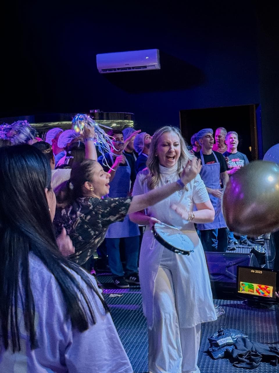 Mulher tocando pandeiro e sorrindo em meio à equipe do evento, em um momento de celebração nos bastidores.